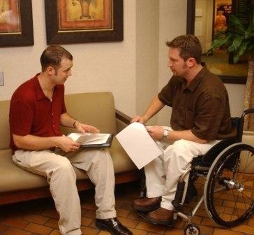 a man on a sofa and a male wheelchair user talking about some papers